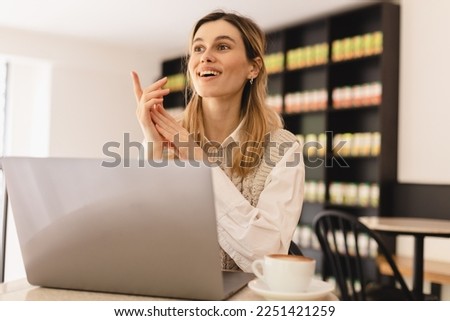 Have idea! Portrait of creative positive attractive young girl freelancer in knitted beige vest and white shirt is sitting in cafe and working on laptop with toothy smile and showing finger up. indoor Royalty-Free Stock Photo #2251421259