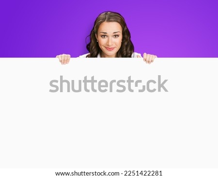 Smiling business woman standing beehind, peeping out white empty mock up signboard. Success and advertising concept. Copy space place, over violet purple background background. Young businesswoman.