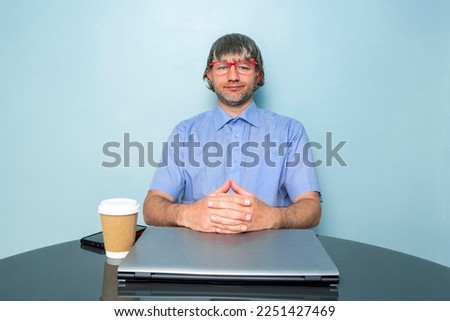 Happy office worker at laptop computer with his phone. Male model wearing blue shirt. Happy face expression. Man in his 40s with short grey beard and grey hairs. Cup of coffee on the table.