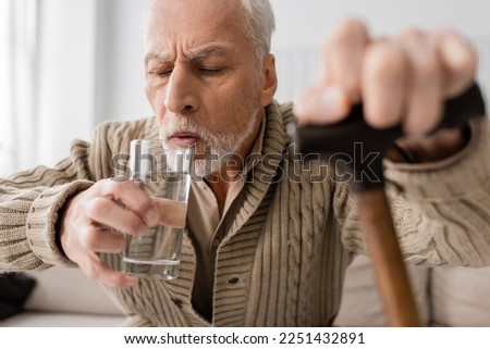 grey haired man with parkinson disease and trembling hands drinking water while sitting with blurred walking cane at home Royalty-Free Stock Photo #2251432891