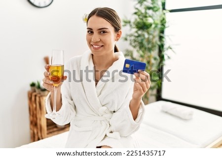 Young hispanic woman holding credit card drinking champagne at beauty center.