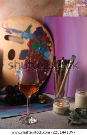 Glass of tasty wine, brushes with colorful paints and burning candle on light gray table