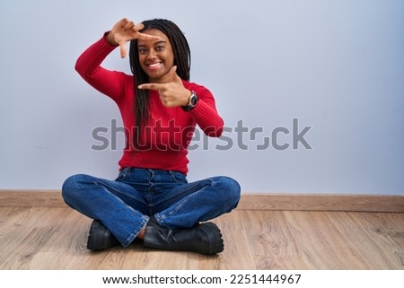 Young african american with braids sitting on the floor at home smiling making frame with hands and fingers with happy face. creativity and photography concept. 