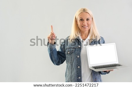 Mature blonde woman with laptop pointing at something on light background