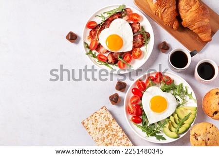 Fried eggs in heart shape, bacon, avocado and vegetables on two white plates, coffee cups and muffins- couple breakfast on Valentines Day or honeymoon on gray background, food border, copy space