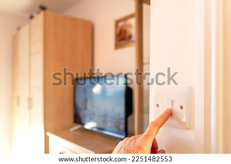 Shallow focus of a homeowner turning on a light switch in bedroom interior. A smart TV and cupboards can be seen in the room. Royalty-Free Stock Photo #2251482553