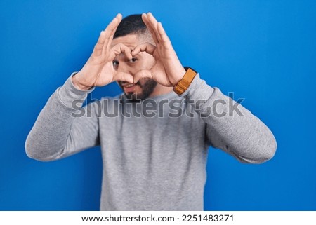 Hispanic man standing over blue background doing heart shape with hand and fingers smiling looking through sign 