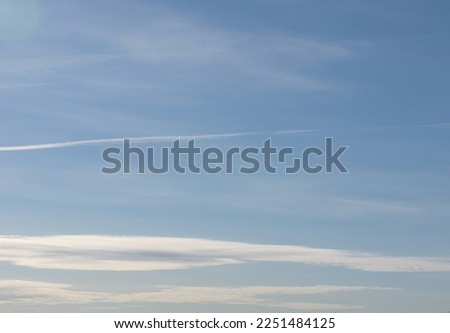 Pale blue sky with pale feathery and bright layered clouds above horizon. Contrail from plane. Gradient of color and illumination.

