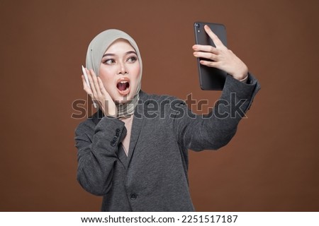 Beautiful Asian hijab influencer in formal office attire smiling and happy taking a selfie with mobile tablet isolated over dark background. Technology, content creator lifestyle concept. 