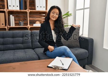 Young asian woman at consultation office smiling cheerful presenting and pointing with palm of hand looking at the camera. 