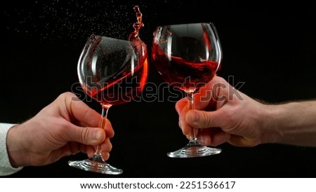Two men clinking with glasses of red wine, celebrating success or speaking toast, close-up. Isolated on black background. Wine is splashing out of glasses. Royalty-Free Stock Photo #2251536617