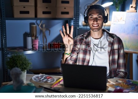 Young hispanic man sitting at art studio with laptop late at night showing and pointing up with fingers number four while smiling confident and happy. 