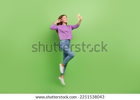 Full size photo of cute positive girl dressed jeans purple pullover hold smartphone showing v-sign isolated on green color background