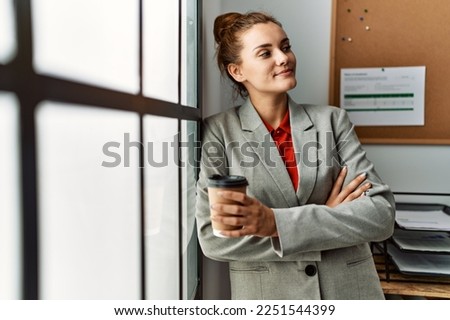Young woman business worker drinking coffee standing with arms crossed gesture at office
