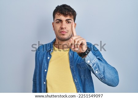 Young hispanic man standing over blue background pointing with finger up and angry expression, showing no gesture 