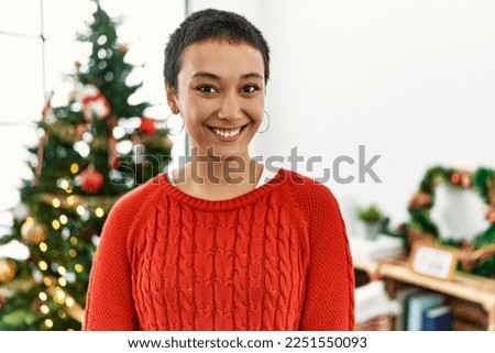 Young hispanic woman smiling confident standing by christmas tree at home