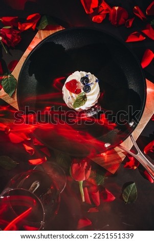 black frying pan with a pavlova cake and scattered red rose petals on a wooden board with a black background with a glass of red wine and lit candles next to it. long exposure with red light. flat lay