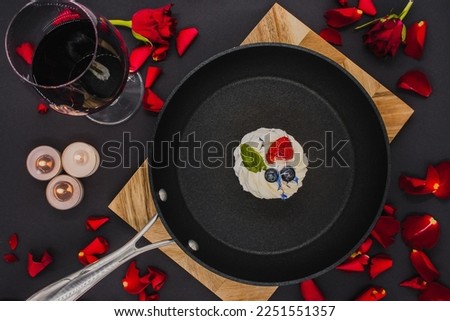 black frying pan with a pavlova cake on a wooden board with a black background, next to which is a glass of red wine and lit candles. flat lay