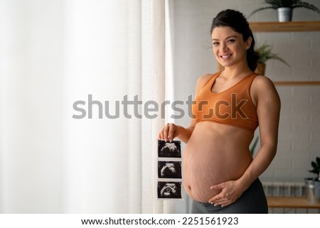 Happy pregnant woman with stretch marks and a beautiful smile in comfortable clothes showing her belly, holding ultrasound pictures in one hand while another hand is on the stomach.