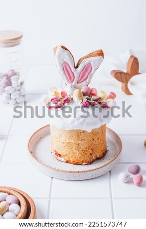 Easter Panettone or Kulich, sweet bread decorated with bunny ears cookies, mini chocolate eggs, marshmallows and dry roses. Kulich on a white background. Royalty-Free Stock Photo #2251573747