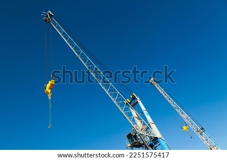 The tops of two-port cranes with a stabilizer and a hook against a blue sky. Royalty-Free Stock Photo #2251575417