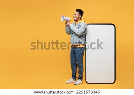 Full body fun young man of African American ethnicity wear denim jacket hoody near big huge blank screen mobile cell phone smartphone with area scream in megaphone isolated on plain yellow background