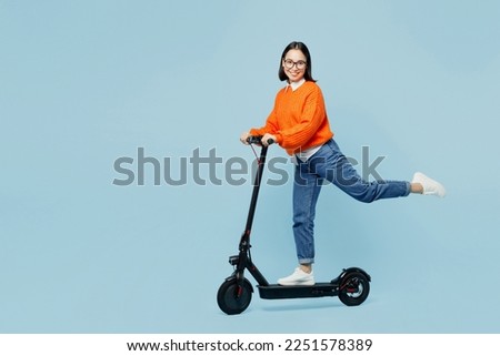 Full body side view smiling happy young woman of Asian ethnicity wear orange sweater glasses riding electric scooter isolated on plain pastel light blue cyan background studio People lifestyle concept Royalty-Free Stock Photo #2251578389