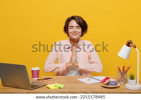 Young employee business woman wear casual shirt sit work at office desk hold hand folded in prayer gesture beg ask about something isolated on plain yellow color background. Achievement career concept