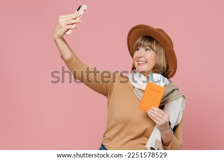 Traveler tourist mature elderly woman 55 years old wear brown shirt hat scarf hold passport boarding tickets do selfie shot on mobile cell phone isolated on plain light pink background studio portrait