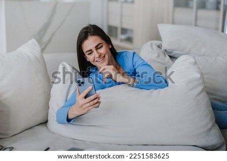 Adorable brunette with  laying on sofa leaning on a big pillow, dressed in blue sweatshirt, makes video call, social media, smiling broadly. Student speaks by phone with parent. Happy people . Blogger