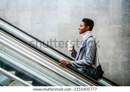 Side view of stylish African American female in stylish coat and sweater. She is with cup of coffee and bag smiling and riding moving stairs in city Royalty-Free Stock Photo #2251605777