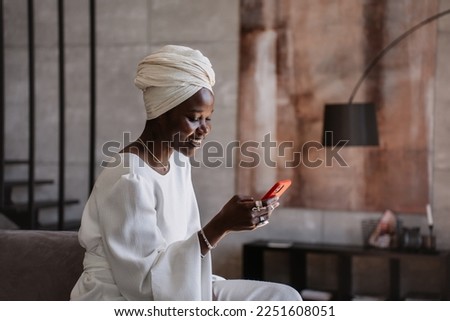 Happy african young woman in white turban and white dress sits on cozy sofa at hotel, reads funny message from friend. Cheerful Brazilian girl surfing social media laughs. Grateful female chatting. Royalty-Free Stock Photo #2251608051