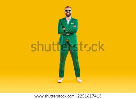 Portrait of cheerful cool and joyful man in green suit and sunglasses of same color. Funny man who celebrates St. Patrick's Day standing with folded hands and smiling at camera on orange background.