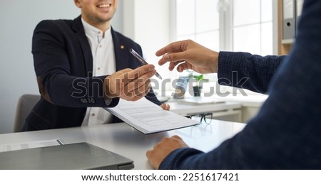 Two young people make a business deal and sign a contract agreement. Smiling agent sitting at his office desk and handing over a pen and a paper document to his client. Cropped shot. Banner background Royalty-Free Stock Photo #2251617421