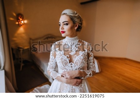 A beautiful blonde bride in a lace white dress with a decollete stands in a room, interior. Wedding photography, close-up portrait.