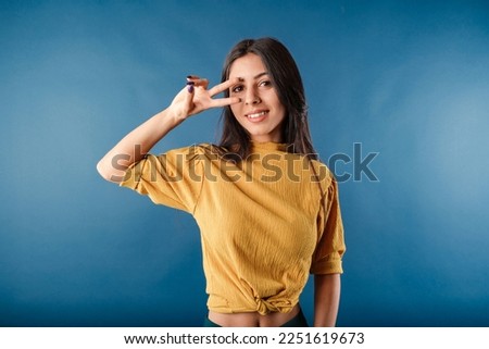 Cute dark-haired woman wearing casual top isolated over blue background doing peace symbol with fingers over face, smiling cheerful showing victory.