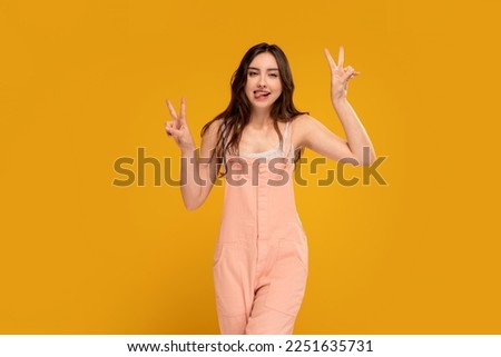 Positive, young, happy woman having fun in studio, sticking tongue and showing peace sign, victory gesture. Yellow background. Copy space. Real people emotions. 
