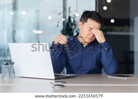 Asian worker tired at work has pain in eyes, businessman in glasses working late, using laptop at work inside office, serious and concentrated pensive. Royalty-Free Stock Photo #2251650179