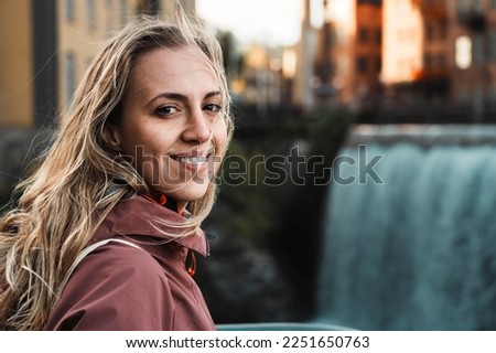 Beautiful girl near a large waterfall. Blonde tourist smiles looking at the camera. Young woman with windblown hair in Norrköping. Enjoyment of traveling and discovering the world. Royalty-Free Stock Photo #2251650763