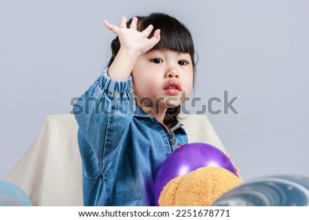Studio shot of little cute preschooler girl daughter in casual street wears jeans jacket and sneakers sitting smiling teddy bear doll on floor playing with colorful plastic balls on gray background