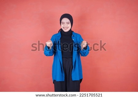 Beautiful Asian woman in blue shirt and hijab smiling cheerful pointing down on brown background