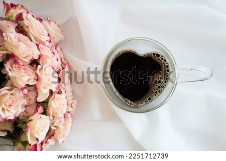 Heart-shaped glass cup filled with coffee on white satin fabric, pink roses on left with pictured, top view, horizontal
