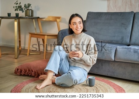 Image of stylish young woman in modern house, using mobile phone, sitting on floor and holding smartphone, drinking from cup. Royalty-Free Stock Photo #2251735343