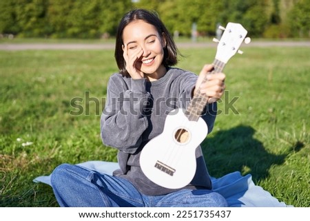 Beautiful asian girl with happy smile, shows her ukulele, sits outside in park on grass, relaxes with music.