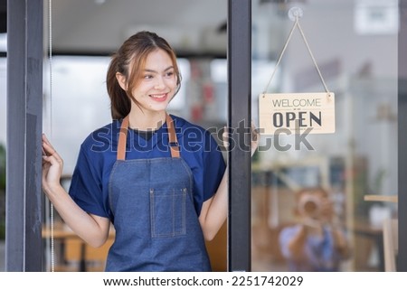 Shot of smiling young cafe show owner Asian woman is a waitress in an apron standing with arms crossed in the doorway waiting for customers Open sign Small business concept, cafes and restaurants.