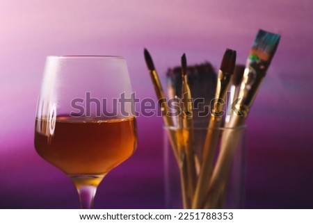 Glass of tasty wine, brushes with colorful paints near gradient canvas, closeup. Space for text