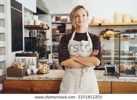 Smiling female baker entrepreneur standing at the counter of bakery and coffee shop. Young woman in cafe near showcase with fresh croissants and bread. Local small business owner indoors. Royalty-Free Stock Photo #2251852201