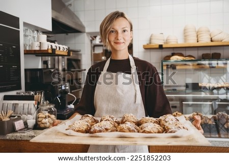Welcoming female baker holding freshly baked almond croissants in  background of bakery. Young smiling seller woman in cafe or coffee shop. Bistro owner indoors. Sustainable local small business. Royalty-Free Stock Photo #2251852203