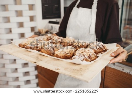 Welcoming female baker holding freshly baked almond croissants in  background of bakery. Young smiling seller woman in cafe or coffee shop. Bistro owner indoors. Sustainable local small business.