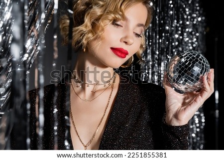 blonde young woman with red lips holding small disco ball near tinsel curtain on grey Royalty-Free Stock Photo #2251855381